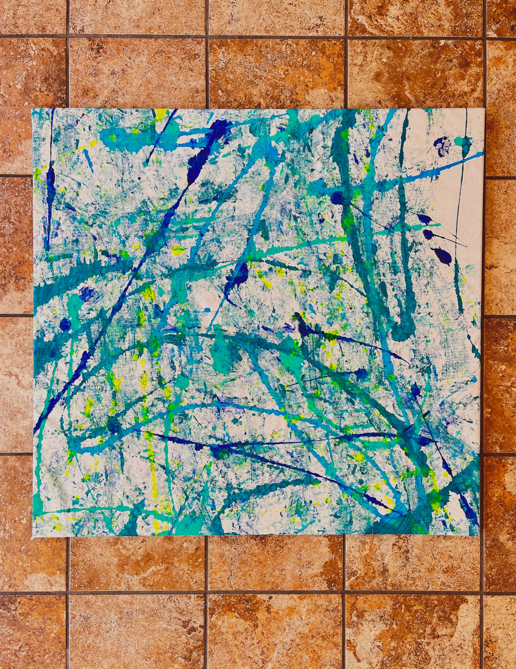 An abstract canvas painting with blue, teal, and yellow paint splatters and drips on a white background, displayed flat on a brown tiled floor.