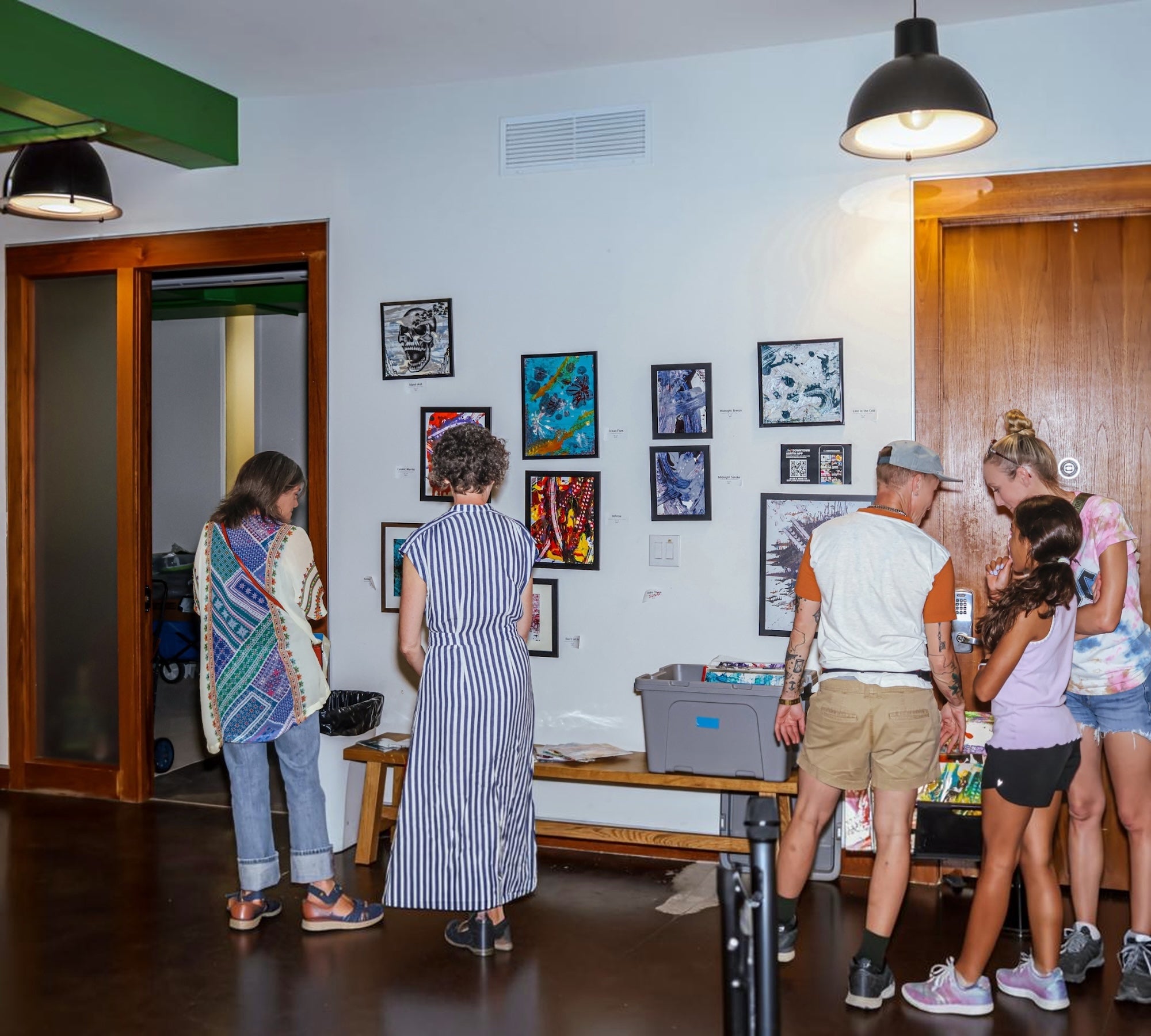 A group of people gathered in a room to view a collection of framed art prints displayed on a white wall. The room features dark wood floors, a wooden door on the right, and a table with storage bins in the foreground.